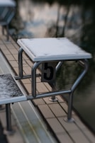 A poolside diving block covered in a thin layer of snow, with the number 5 clearly visible. The surface is slick and white, indicating cold weather. The water in the background is calm and dark, reflecting the sky.
