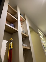 A warmly lit modern interior with wooden shelves set into a wall. The shelves hold various decorative items, including a figurine of a person dressed in vibrant yellow, blue, and red colors, a small bunch of purple flowers, books, and other small decorations. The overall decor blends natural elements with contemporary design.