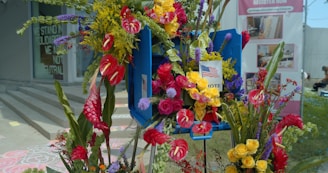 A vibrant floral arrangement is prominently displayed, featuring a variety of colorful flowers such as red anthuriums, yellow roses, and purple accents. An American flag and a sign with the word VOTE are nestled among the blooms. In the background, there are signs advocating solidarity and promoting voter registration, set against the backdrop of a building and a grassy area.