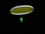 A badminton racket is suspended in the air against a black background. The strings of the racket are illuminated in a bright lime green color, contrasting with the dark surroundings. The handle of the racket is partially visible in a blurred focus.