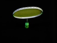 A badminton racket is suspended in the air against a black background. The strings of the racket are illuminated in a bright lime green color, contrasting with the dark surroundings. The handle of the racket is partially visible in a blurred focus.
