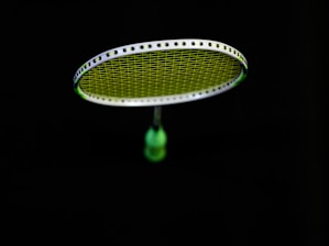 A badminton racket is suspended in the air against a black background. The strings of the racket are illuminated in a bright lime green color, contrasting with the dark surroundings. The handle of the racket is partially visible in a blurred focus.
