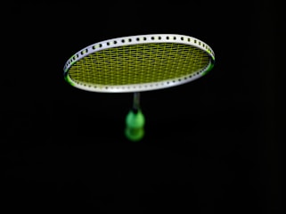 A badminton racket is suspended in the air against a black background. The strings of the racket are illuminated in a bright lime green color, contrasting with the dark surroundings. The handle of the racket is partially visible in a blurred focus.