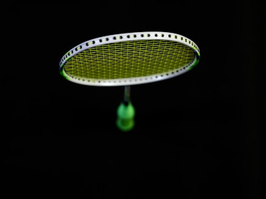 A badminton racket is suspended in the air against a black background. The strings of the racket are illuminated in a bright lime green color, contrasting with the dark surroundings. The handle of the racket is partially visible in a blurred focus.