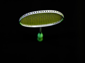 A badminton racket is suspended in the air against a black background. The strings of the racket are illuminated in a bright lime green color, contrasting with the dark surroundings. The handle of the racket is partially visible in a blurred focus.
