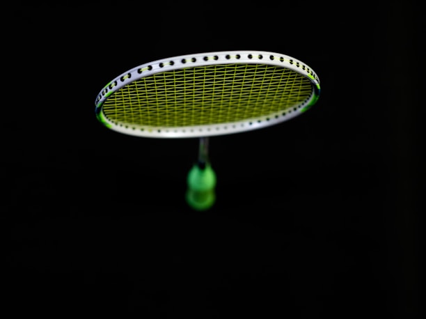 A badminton racket is suspended in the air against a black background. The strings of the racket are illuminated in a bright lime green color, contrasting with the dark surroundings. The handle of the racket is partially visible in a blurred focus.