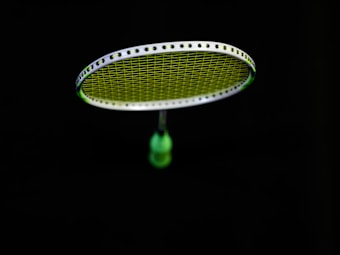 A badminton racket is suspended in the air against a black background. The strings of the racket are illuminated in a bright lime green color, contrasting with the dark surroundings. The handle of the racket is partially visible in a blurred focus.