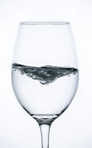 A clear glass filled with water, featuring bubbles and ripples on the surface. The glass is positioned against a white background, highlighting the transparency and reflective properties of the glass and the fluid inside.