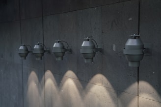 A row of five industrial-style wall-mounted light fixtures is evenly spaced along a textured concrete wall. Each fixture casts a downward beam of light, creating a pattern of illuminated spots on the surface below.