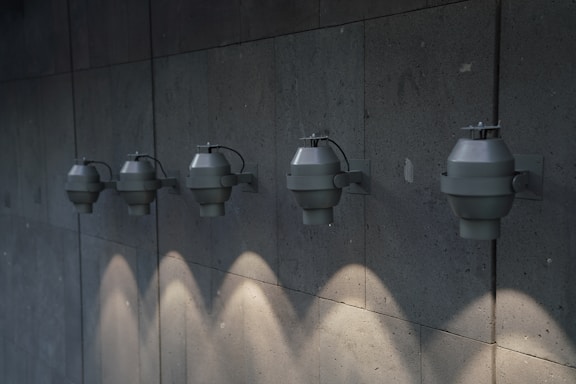 A row of five industrial-style wall-mounted light fixtures is evenly spaced along a textured concrete wall. Each fixture casts a downward beam of light, creating a pattern of illuminated spots on the surface below.