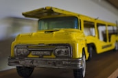 A vintage toy truck with a bright yellow paint job and metallic gray details is positioned on a wooden surface. The toy appears to be well-worn, showing signs of age and use.
