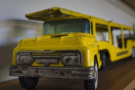 A vintage toy truck with a bright yellow paint job and metallic gray details is positioned on a wooden surface. The toy appears to be well-worn, showing signs of age and use.