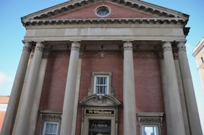A classical building with a red brick facade features four large stone columns with Corinthian capitals. The building has an ornate triangular pediment and decorative elements above the doorway, which displays a sign reading 'The Windamere, 2 S. Main St.' Windows are symmetrically placed on both sides.