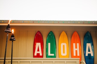 Five colorful surfboards are displayed vertically against a wall with the letters spelling 'ALOHA'. Two tiki torches are lit in the foreground, adding a warm, tropical vibe. Multicolored string lights are hung along the roofline above the surfboards.