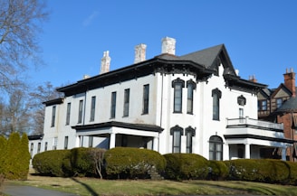 A large, two-story, white historical house with ornate black window frames and several chimneys. The house is surrounded by neatly trimmed bushes and trees, set against a clear blue sky. The architecture features elements typical of older, classic styles, with a prominent roof and a balanced, symmetrical design.
