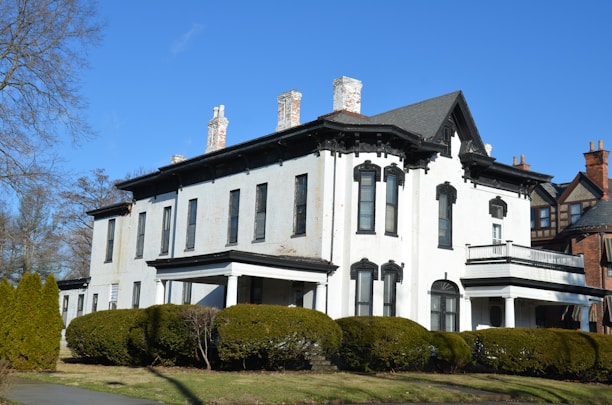 A large, two-story, white historical house with ornate black window frames and several chimneys. The house is surrounded by neatly trimmed bushes and trees, set against a clear blue sky. The architecture features elements typical of older, classic styles, with a prominent roof and a balanced, symmetrical design.