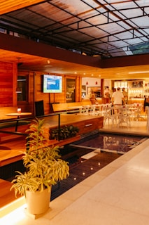 A modern interior space with wooden accents and a high ceiling. There is a large television mounted on the wall displaying a screen. The room features a cozy seating area with white chairs and a bar in the background illuminated by warm lighting. Indoor plants and a small pool add to the calming atmosphere. Several people are casually gathered near the bar.
