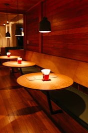 A cozy cafe setting with warm wooden interiors and dim lighting. The seating area consists of round wooden tables with cushioned bench seating along the wall. Black pendant lights hang from the ceiling, casting a soft glow. A red mug with napkins sits on each table, accompanied by a menu or paper.
