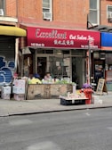 A street view featuring a brick-walled building with a salon named 'Excellent Cut Salon Inc.' The salon has a red awning with both English and Chinese text. In front of the salon, there is a small vegetable and fruit stand with a person sitting behind it. Various produce items including cucumbers, cauliflower, and onions are displayed. Some graffiti can be seen on the wooden and metal surfaces near the stand.