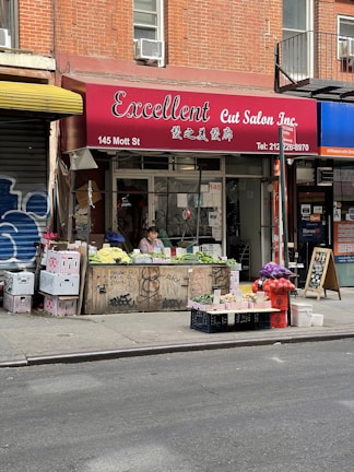 A street view featuring a brick-walled building with a salon named 'Excellent Cut Salon Inc.' The salon has a red awning with both English and Chinese text. In front of the salon, there is a small vegetable and fruit stand with a person sitting behind it. Various produce items including cucumbers, cauliflower, and onions are displayed. Some graffiti can be seen on the wooden and metal surfaces near the stand.