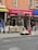 A street view featuring a brick-walled building with a salon named 'Excellent Cut Salon Inc.' The salon has a red awning with both English and Chinese text. In front of the salon, there is a small vegetable and fruit stand with a person sitting behind it. Various produce items including cucumbers, cauliflower, and onions are displayed. Some graffiti can be seen on the wooden and metal surfaces near the stand.