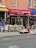 A street view featuring a brick-walled building with a salon named 'Excellent Cut Salon Inc.' The salon has a red awning with both English and Chinese text. In front of the salon, there is a small vegetable and fruit stand with a person sitting behind it. Various produce items including cucumbers, cauliflower, and onions are displayed. Some graffiti can be seen on the wooden and metal surfaces near the stand.
