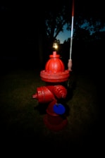 A bright red fire hydrant stands on a grassy area with a dark, shadowy background. Its bold color contrasts sharply with the dim surroundings, and a small reflective disc is attached to it.