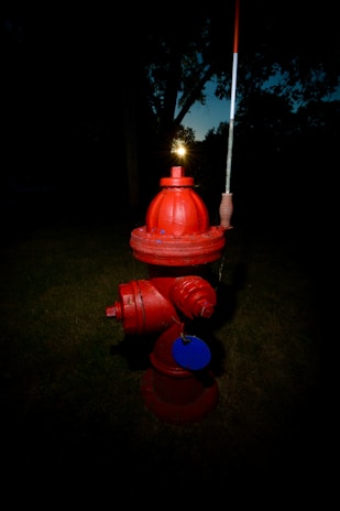A bright red fire hydrant stands on a grassy area with a dark, shadowy background. Its bold color contrasts sharply with the dim surroundings, and a small reflective disc is attached to it.