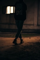 A person wearing dark clothing stands with crossed legs in a dimly lit space. The light outside the small window in the background casts a faint glow. The atmosphere appears somber and mysterious.