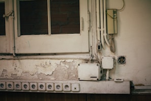 A section of an old wall with peeling paint and multiple electrical outlets. Several yellowed electrical boxes and cables are exposed. The top left shows a wooden window with glass panes, partially revealing brickwork behind.