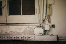 A section of an old wall with peeling paint and multiple electrical outlets. Several yellowed electrical boxes and cables are exposed. The top left shows a wooden window with glass panes, partially revealing brickwork behind.