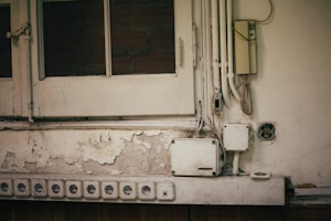 A section of an old wall with peeling paint and multiple electrical outlets. Several yellowed electrical boxes and cables are exposed. The top left shows a wooden window with glass panes, partially revealing brickwork behind.