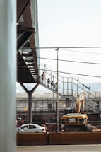 A construction site with a large Caterpillar excavator is present. Above the machinery, a pedestrian bridge made of metal and glass is visible, with people walking along it. In the background, there are cars on a road and industrial structures.