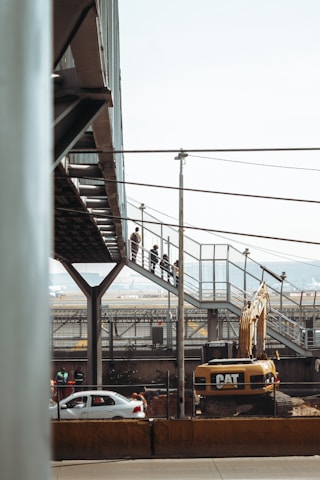 A construction site with a large Caterpillar excavator is present. Above the machinery, a pedestrian bridge made of metal and glass is visible, with people walking along it. In the background, there are cars on a road and industrial structures.