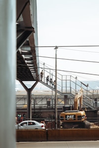 A construction site with a large Caterpillar excavator is present. Above the machinery, a pedestrian bridge made of metal and glass is visible, with people walking along it. In the background, there are cars on a road and industrial structures.