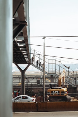 A construction site with a large Caterpillar excavator is present. Above the machinery, a pedestrian bridge made of metal and glass is visible, with people walking along it. In the background, there are cars on a road and industrial structures.