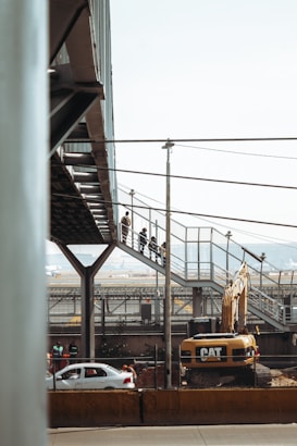 A construction site with a large Caterpillar excavator is present. Above the machinery, a pedestrian bridge made of metal and glass is visible, with people walking along it. In the background, there are cars on a road and industrial structures.