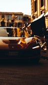 A sleek, modern sports car with a shiny, reflective surface is parked on a cobblestone street. Buildings and various film production equipment can be seen in the background. The image has a warm, golden hue, giving it a cinematic and luxurious feel.