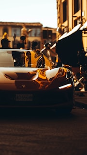 A sleek, modern sports car with a shiny, reflective surface is parked on a cobblestone street. Buildings and various film production equipment can be seen in the background. The image has a warm, golden hue, giving it a cinematic and luxurious feel.