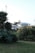 A serene rural scene featuring two houses surrounded by lush greenery. Tall trees and various shrubs fill the foreground, creating a natural frame for the blue-grey houses in the background, which appear to be bathed in soft, natural light.