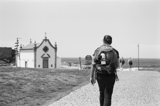A black and white photograph of a person with a backpack walking on a cobblestone path towards a small church near the sea. Additional people are seen walking ahead on the right side towards the water. The scene has a serene and contemplative feel with the ocean in the background and some grassy areas adjacent to the path.