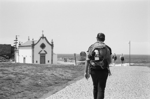 A black and white photograph of a person with a backpack walking on a cobblestone path towards a small church near the sea. Additional people are seen walking ahead on the right side towards the water. The scene has a serene and contemplative feel with the ocean in the background and some grassy areas adjacent to the path.