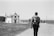 A black and white photograph of a person with a backpack walking on a cobblestone path towards a small church near the sea. Additional people are seen walking ahead on the right side towards the water. The scene has a serene and contemplative feel with the ocean in the background and some grassy areas adjacent to the path.
