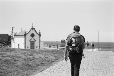 A black and white photograph of a person with a backpack walking on a cobblestone path towards a small church near the sea. Additional people are seen walking ahead on the right side towards the water. The scene has a serene and contemplative feel with the ocean in the background and some grassy areas adjacent to the path.