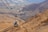 A cable car hanging from cables traverses a rocky, arid landscape with rugged mountains and sparse vegetation. The terrain is predominantly sandy with hues of tan and brown. In the background, there are structures, possibly part of a settlement or tourist facilities, amidst the desert.
