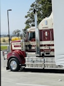 A large red and white semi-truck is parked in a lot, with branding and decoration visible on its side. The tractor features cylindrical exhaust pipes and polished chrome elements. Trees and distant fields can be seen in the background.