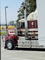 A large red and white semi-truck is parked in a lot, with branding and decoration visible on its side. The tractor features cylindrical exhaust pipes and polished chrome elements. Trees and distant fields can be seen in the background.