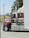 A large red and white semi-truck is parked in a lot, with branding and decoration visible on its side. The tractor features cylindrical exhaust pipes and polished chrome elements. Trees and distant fields can be seen in the background.