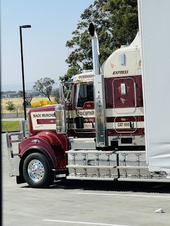 A large red and white semi-truck is parked in a lot, with branding and decoration visible on its side. The tractor features cylindrical exhaust pipes and polished chrome elements. Trees and distant fields can be seen in the background.