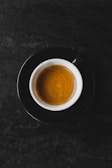 A cup of coffee served in a white cup with a black saucer, placed against a dark textured background. The coffee appears rich and smooth, with a layer of crema on top.